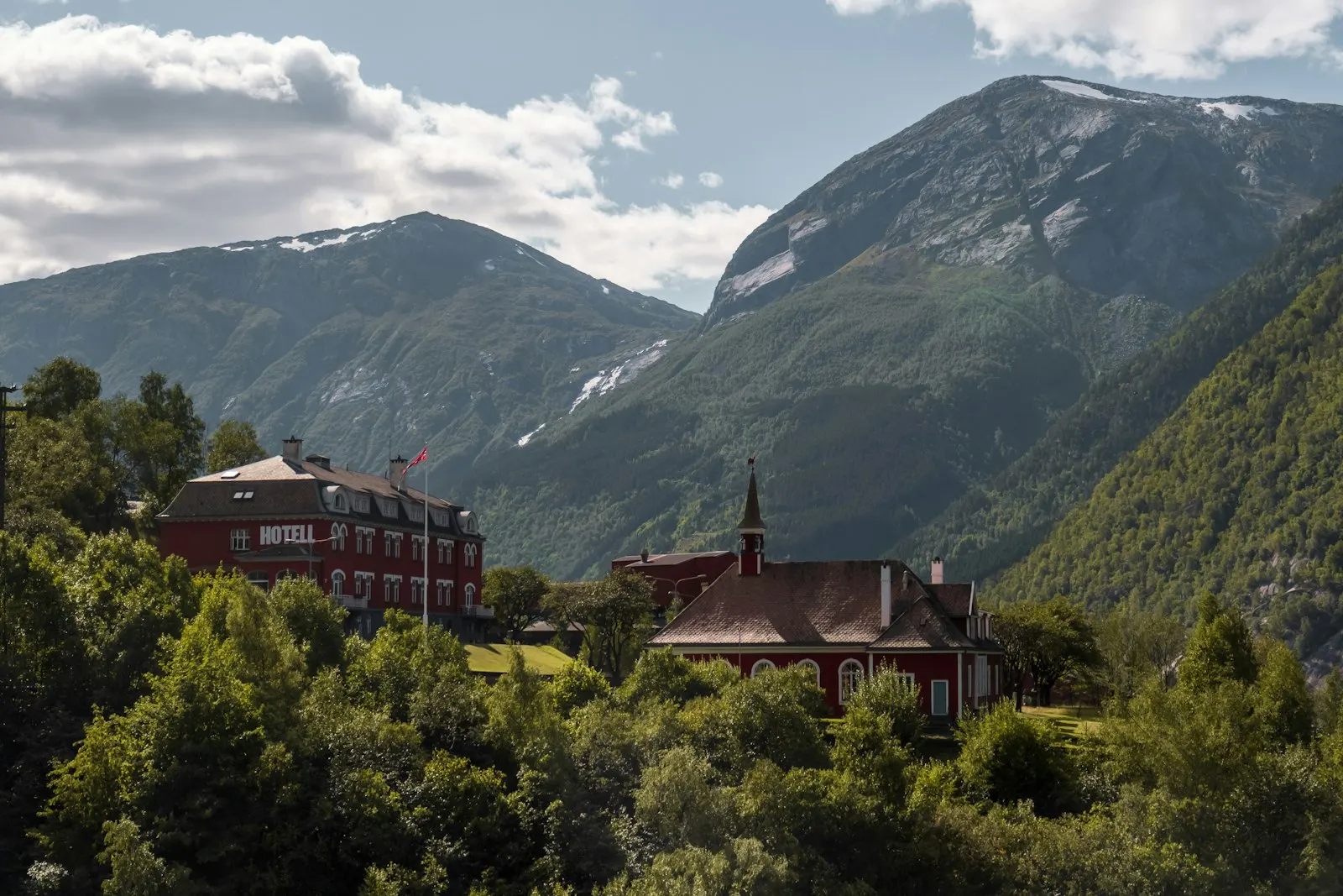 Et rødt, klassisk hotell i tre med norsk flagg, omgitt av sommergrønn skog og fjell i Hardanger